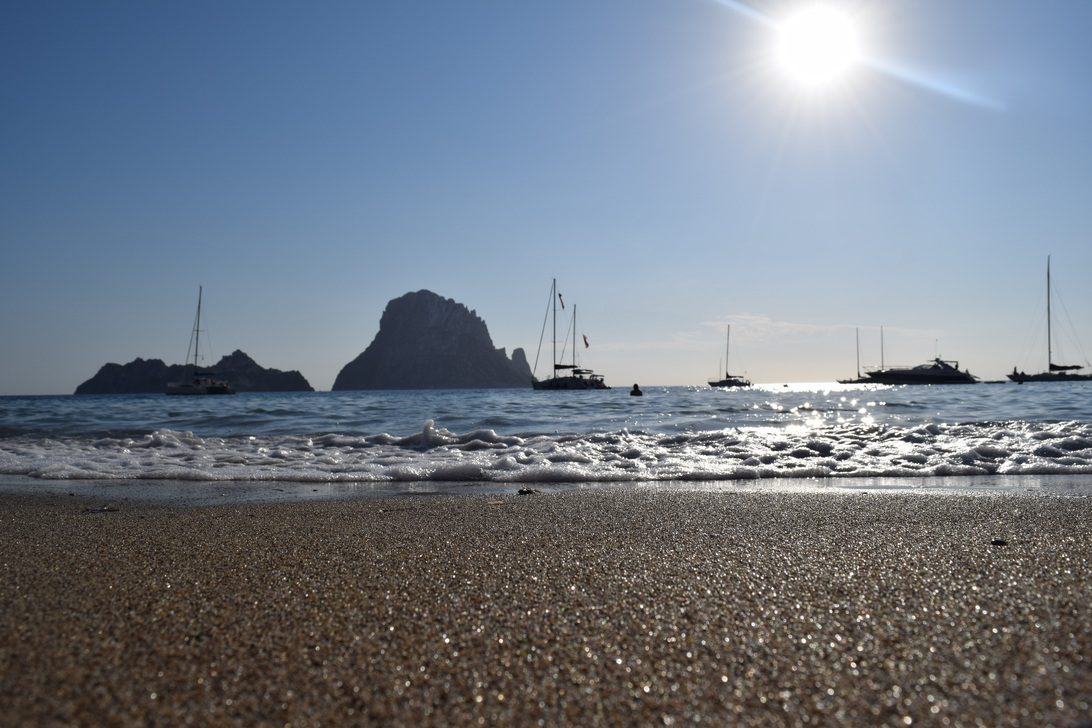 762807 - Play Hotel Ibiza - Pulso Turistico Sunny beach scene with calm sea, sailboats anchored near a distant rocky island under a bright sun to the right.