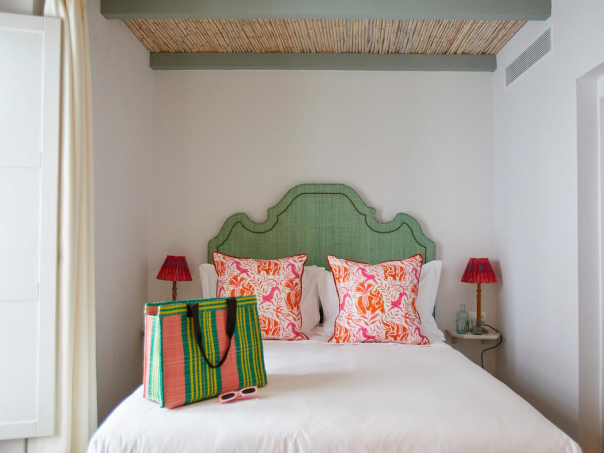 El Escondite - IP - Pulso Turistico Bed with a green upholstered headboard and two orange tropical-pattern pillows, flanked by red lamps and a colorful striped tote on white bedding.