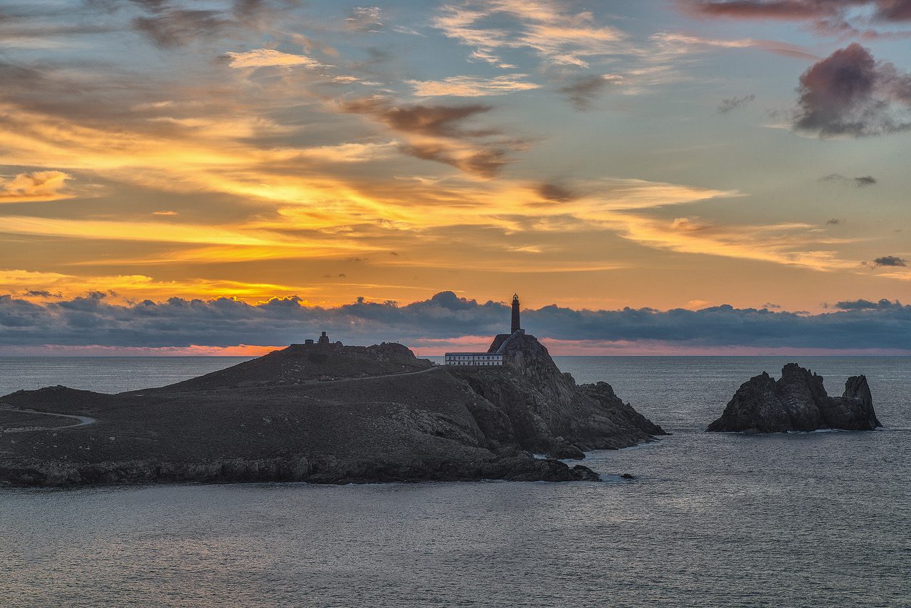 Faro Vilan - A Coruna - IP - Pulso Turistico Lighthouse on a rocky island at sunset with a calm sea and orange-pink sky behind it.”?