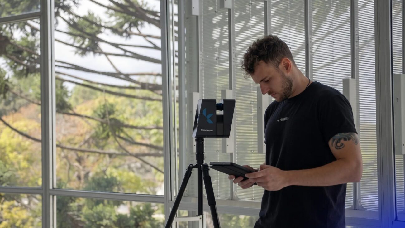 Man in a black t-shirt uses a tablet beside a Matterport 3D scanner on a tripod in a sunlit office with large windows.
