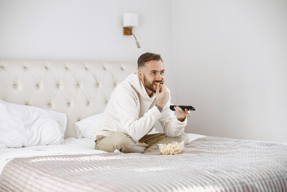 Los televisores inteligentes cambian la experiencia de los huéspedes en hoteles - 780162 - Conekta - Pulso Turistico Los televisores inteligentes cambian la experiencia de los huéspedes en hoteles - Man in a light hoodie sits cross-legged on a bed, eating popcorn and holding a remote while watching TV in a bright room with a tufted headboard.